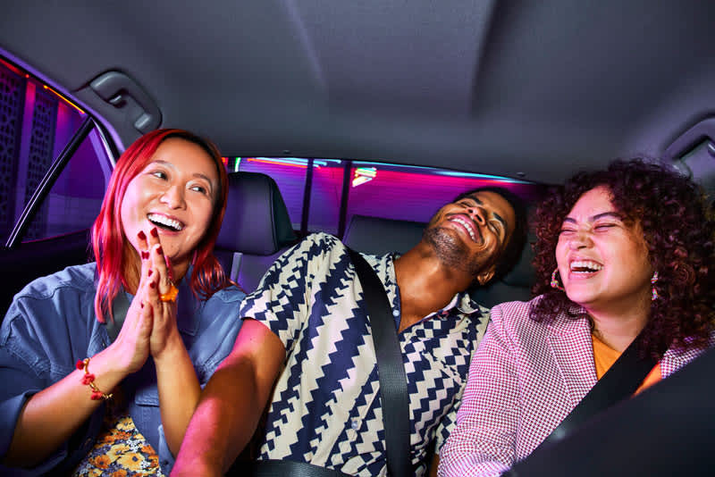 image of two women and man sitting in the back seat of a car smiling and laughing together. 