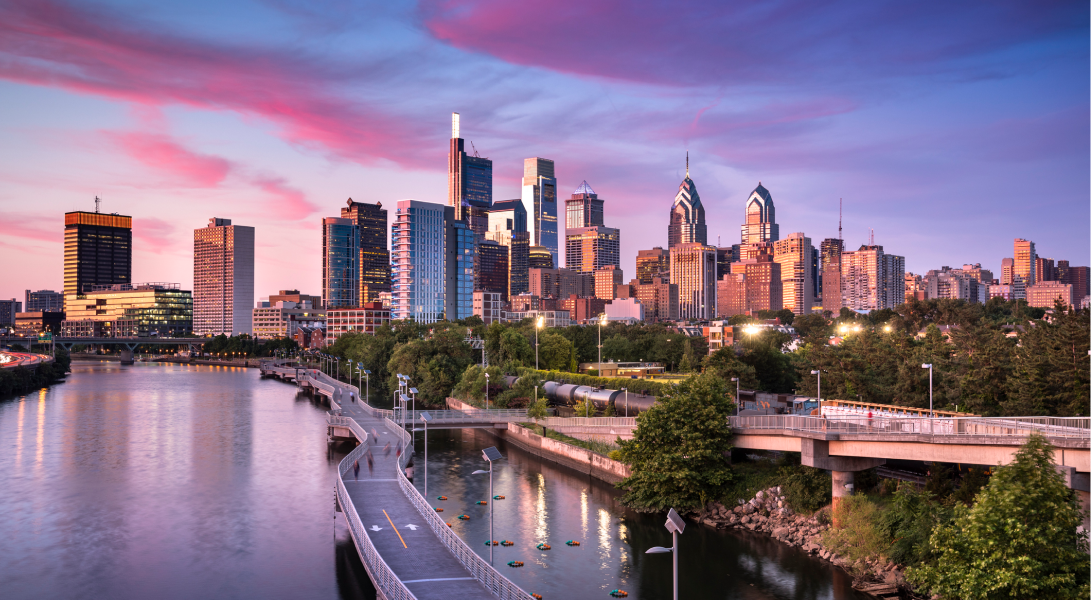 Philadelphia skyline at sunset with tall buildings, river, and pink clouds in the sky.