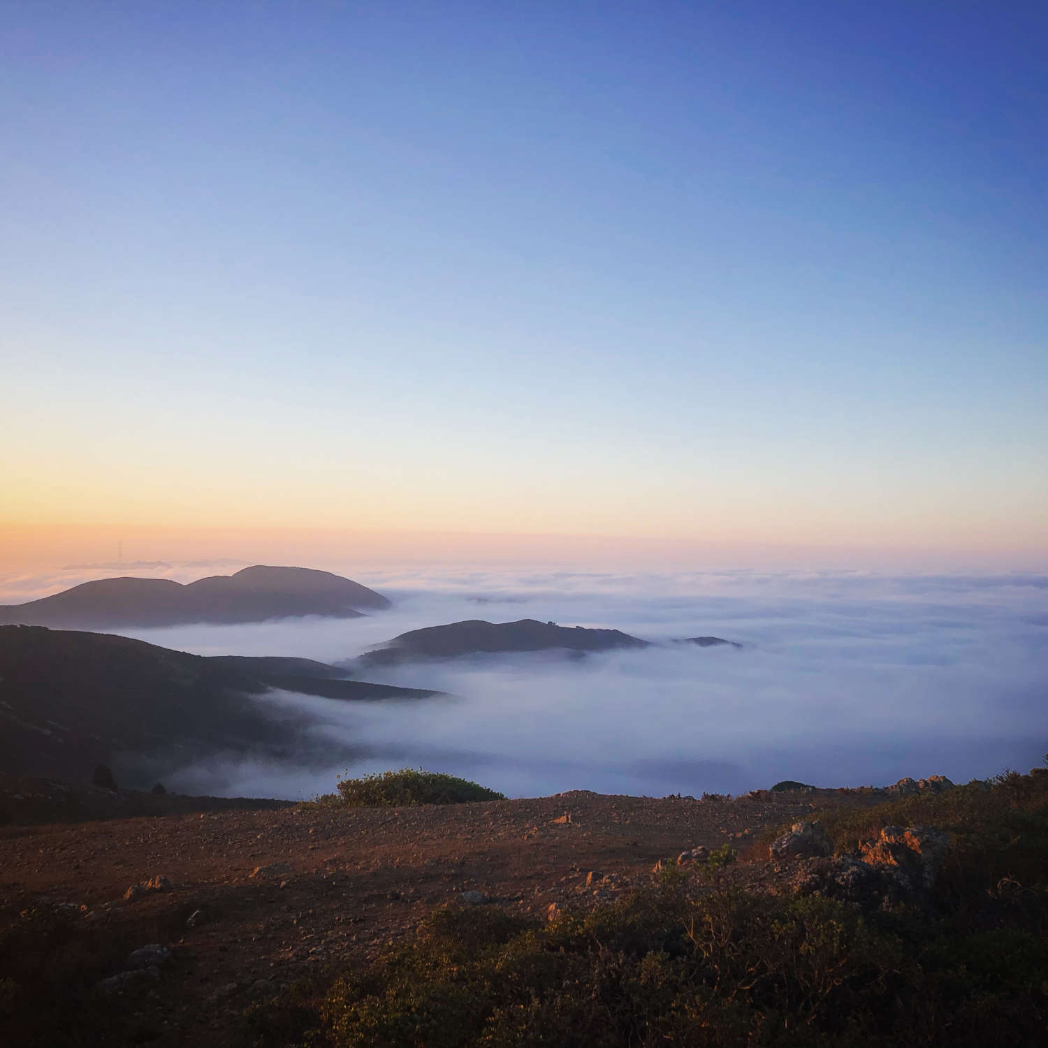 A photo taken by Amy while hiking in the Marin headlands, picturing majestic brown mountain-tops peek out of a layer of clouds, below an orange and blue sky at dusk.