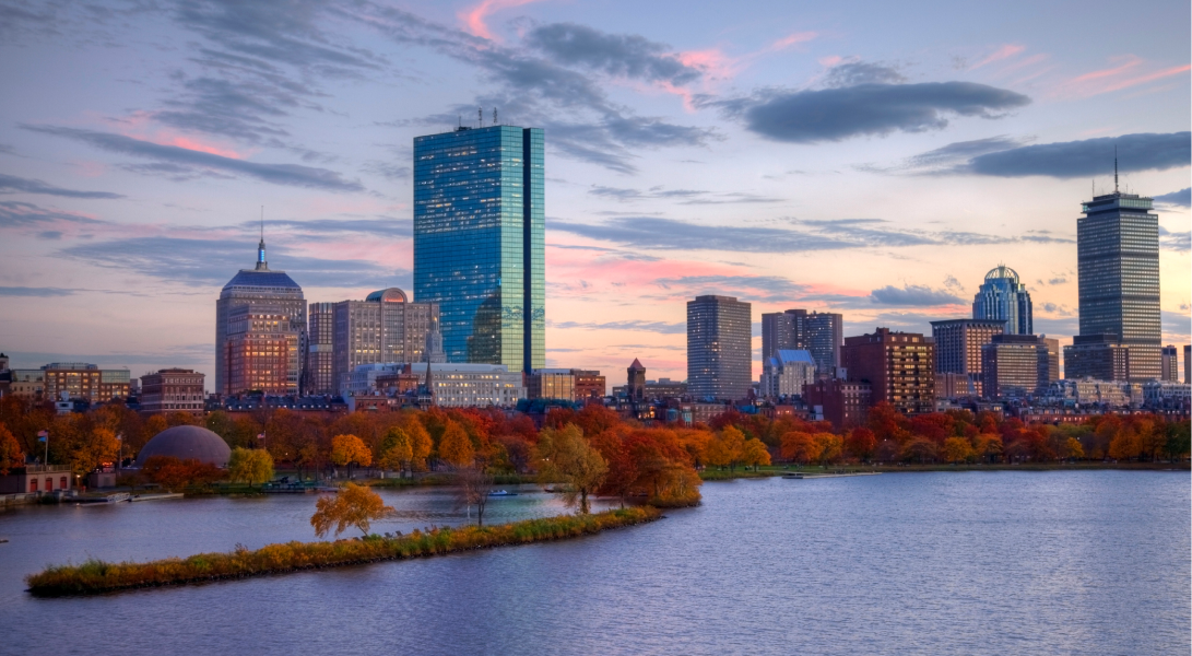 Boston city skyline with tall buildings, autumn trees, and a river at sunset under a colorful sky.