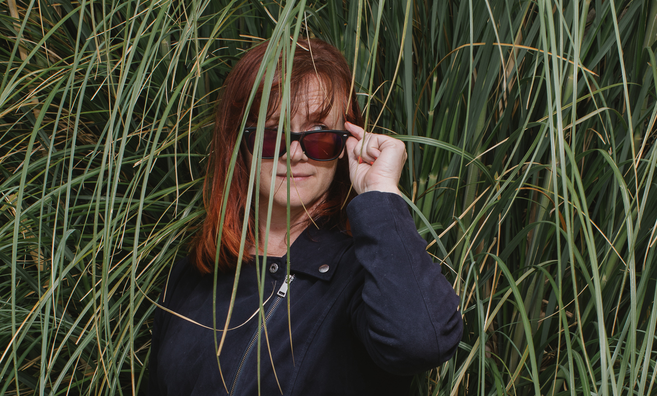 Kat, a woman with light skin and shoulder-length red hair, wears a stylish black jacket and peers sideways over her sunglasses. Behind her is a background of giant grasses.