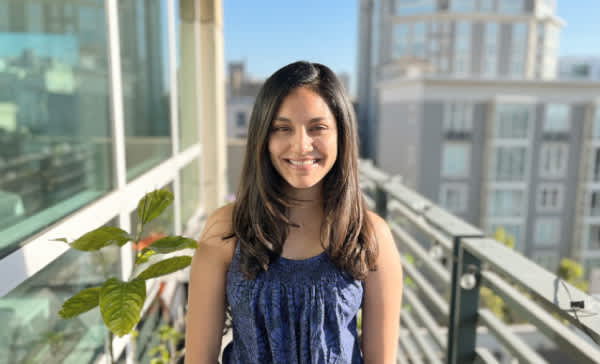 Aastha, a young woman with shoulder-length brown hair and light brown skin, wearing a sleeveless purple top. She stands on a balcony in San Francisco with a city view behind her. 