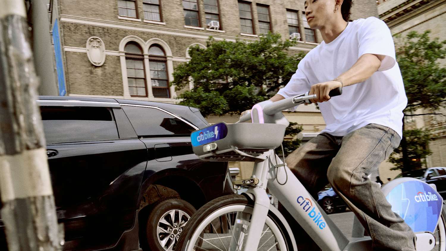 A person in a white shirt rides a Citi Bike past a black minivan on a city street with trees.