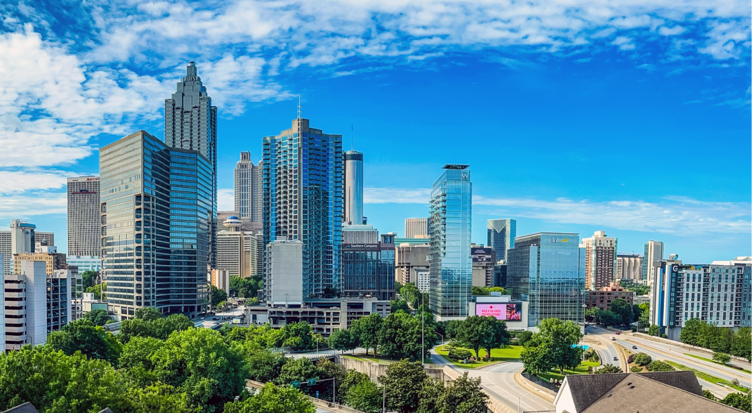 Downtown Atlanta skyline with tall glass buildings, green trees, and a bright blue sky with clouds.