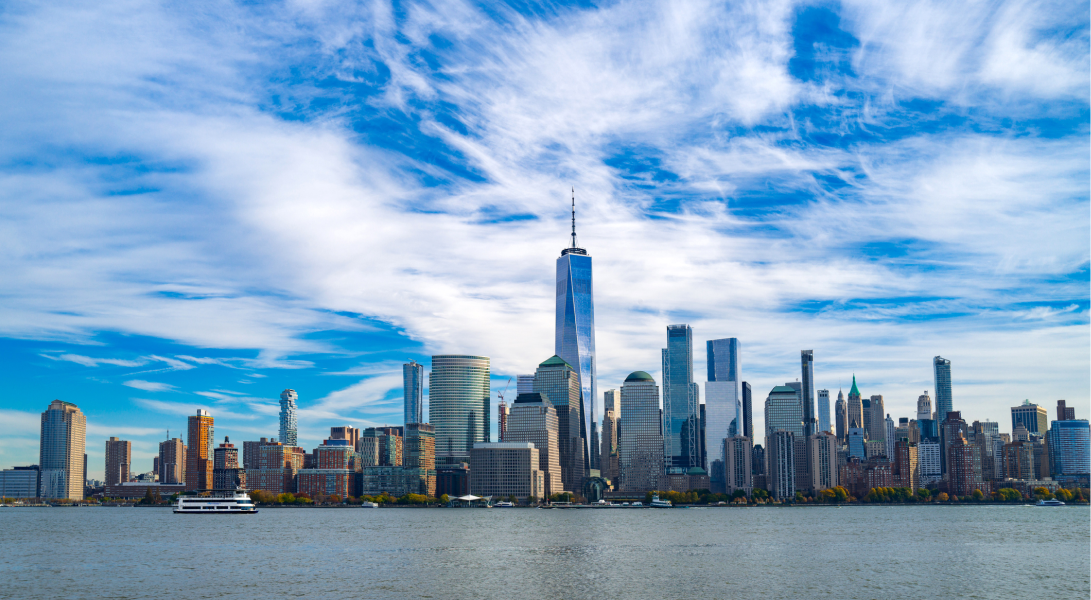 New York City skyline with tall skyscrapers, blue sky, white clouds, and a boat on the river.