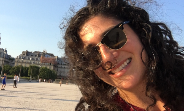 Portrait of Amy Lipner, a woman with sunglasses on, with long brown curly hair standing in a courtyard outside. 