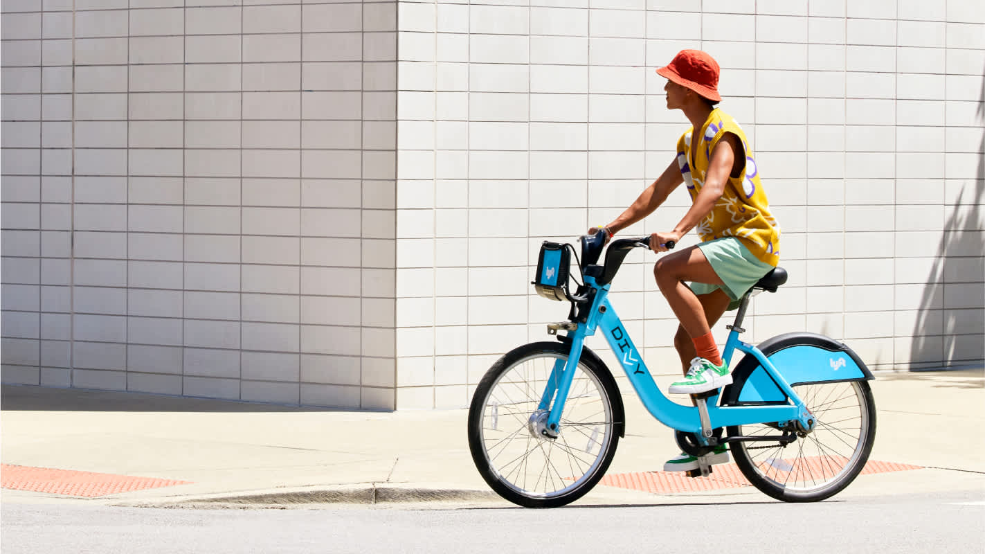 Person in a red hat rides a blue Divvy bike past a white tiled wall on a sunny day.