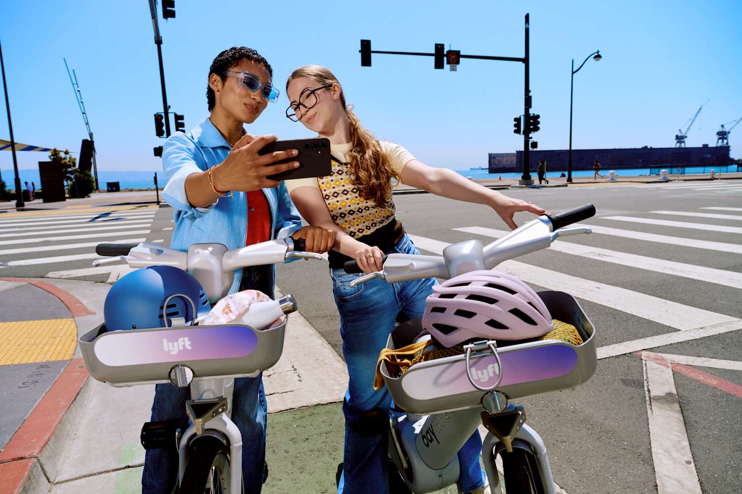 Two people take a selfie with Lyft bikes at a sunny city intersection near the water.