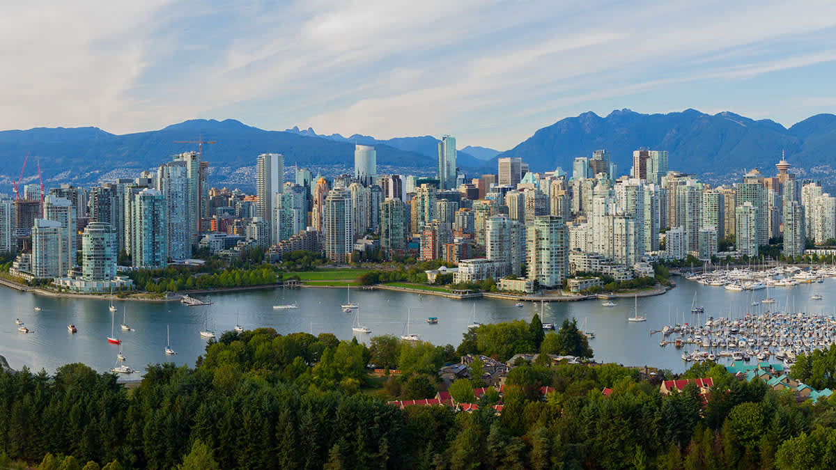 Vancouver city skyline with tall glass buildings, boats in a harbor, green trees, and mountains in the background.