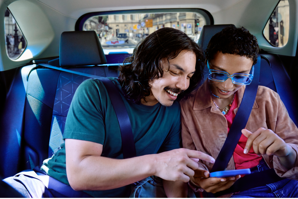 Two friends in a car’s back seat, smiling and looking at a phone, seat belts on
