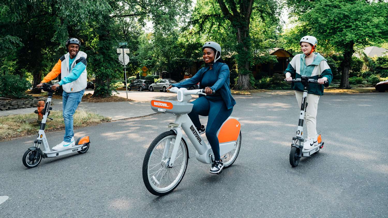 Three people wearing helmets riding BIKETOWN scooters and a BIKETOWN bike on a tree-lined street in daylight.