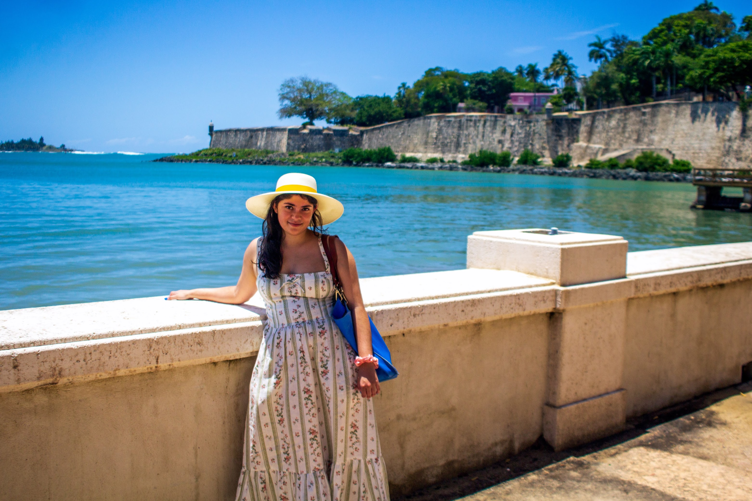 Runi, a young woman with light brown skin and shoulder-length brown hair, wearing a short-sleeved top with diagonal blue stripes. Behind her are water, rocks, and shoreline.