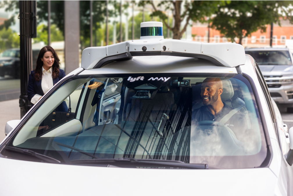 Woman approaches white self-driving Lyft car with a smiling man in the driver's seat on a city street.