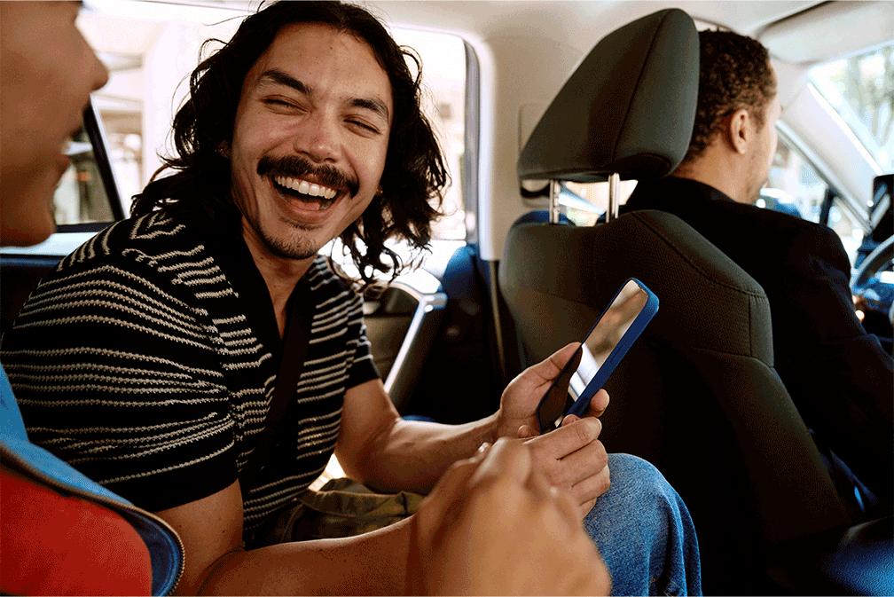 Smiling man with long hair holding a phone, sitting in the backseat of a car with another passenger and a driver in the driver's seat.