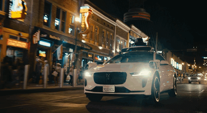 A white SUV with a roof rack is parked on a city street at night in front of a building with glowing signs.