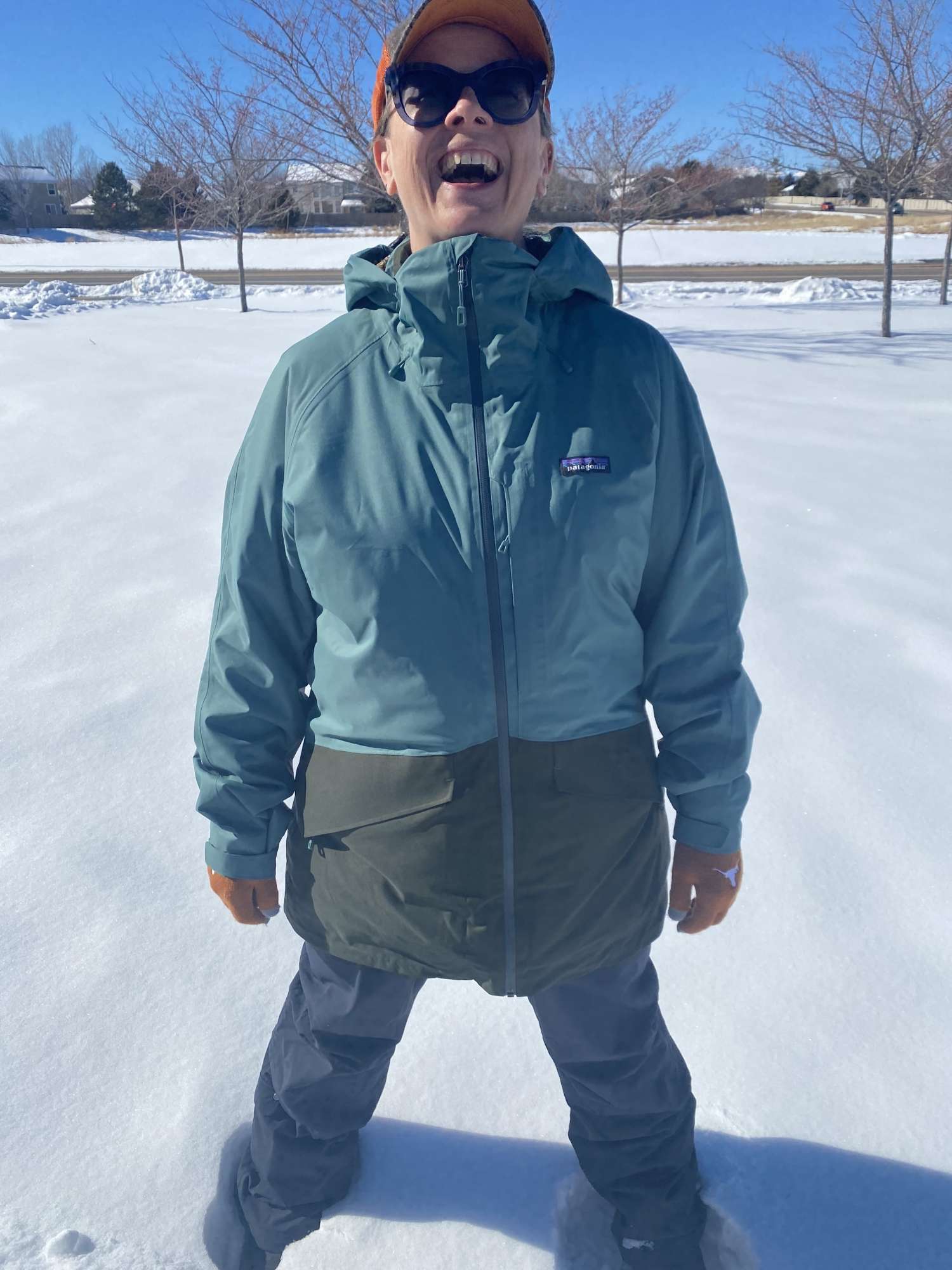 Kat laughing and enjoying the cold outdoors on a sunny day. She wears a baseball hat, sunglasses, blue jacket, and is standing up to her knees in bright white snow.