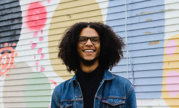 Jacob, a young man with curly brown hair and medium-brown skin, wearing black-rimmed glasses and a denim jacket over a black shirt. Behind him is a mural of colorful circles and shapes.