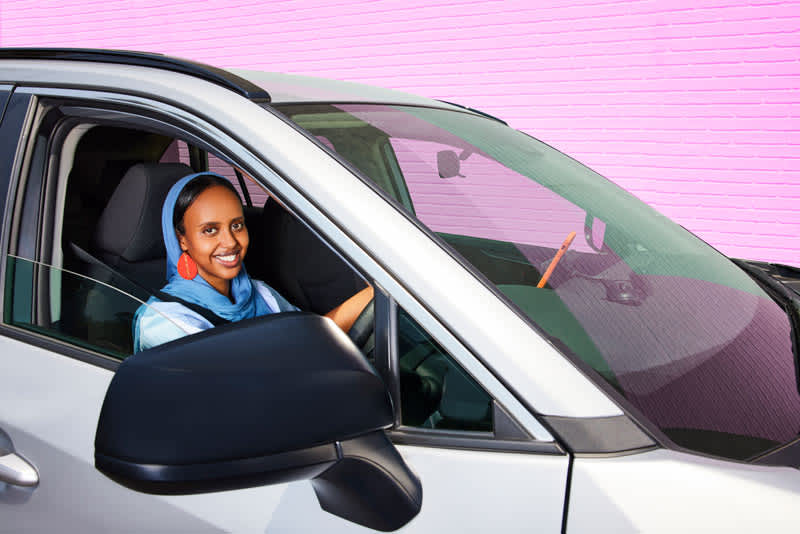 image of a woman wearing a head scarf smiling in the driver seat of a car with a pink colored brick wall in the background. 