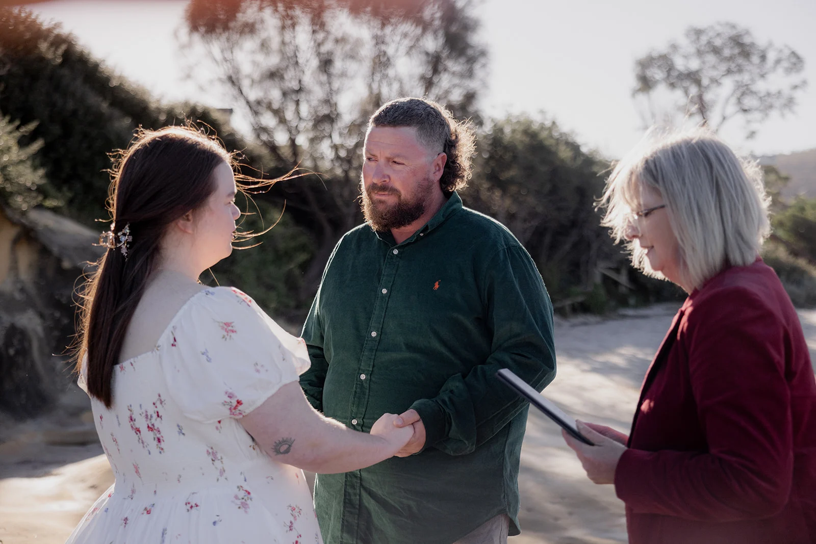 A couple holding hands during an intimate beach elopement ceremony, celebrant Mary Figg reading nearby