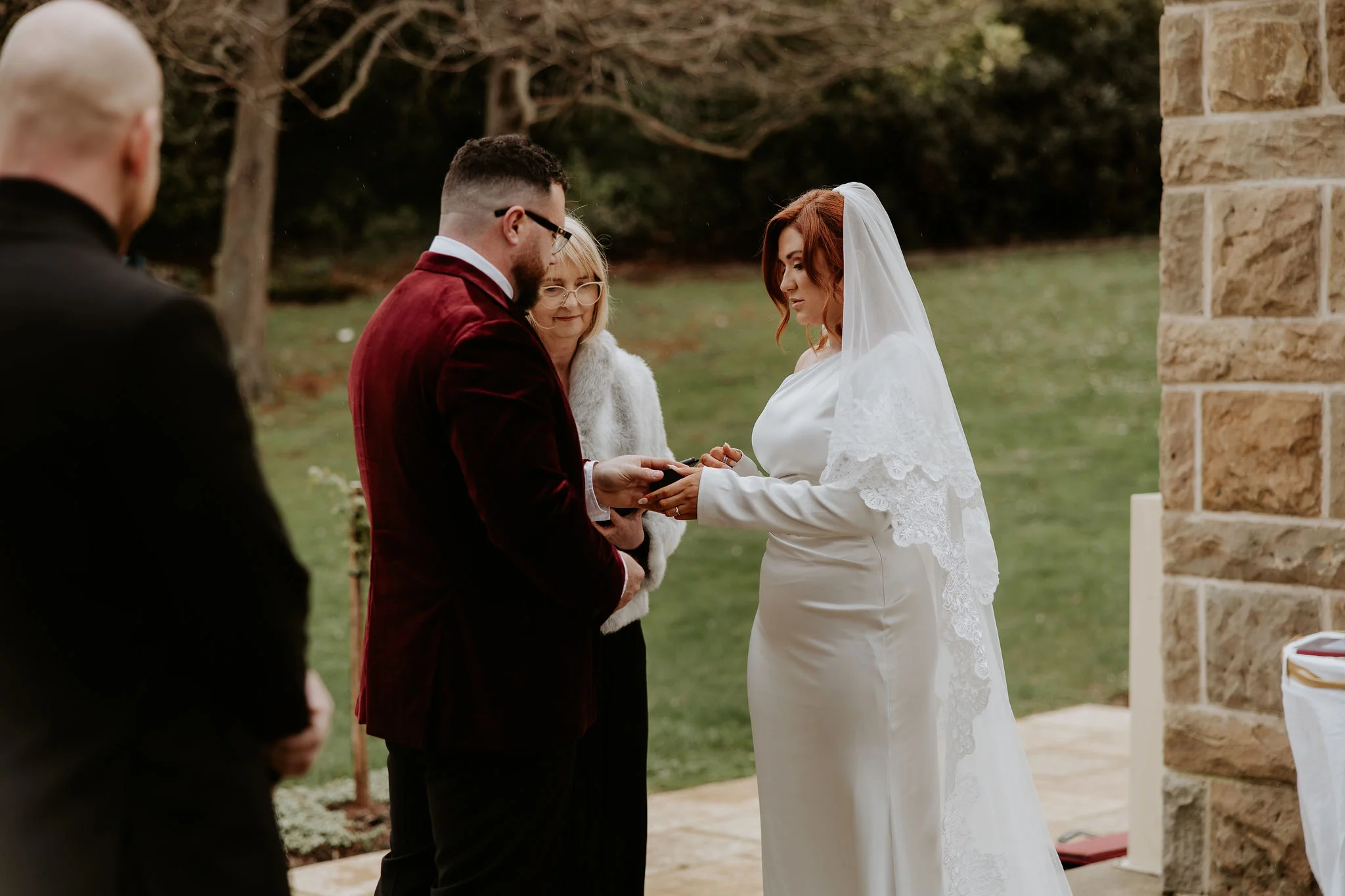 A couple exchanging rings outdoors in winter, the groom's mother looking on in a fur-trimmed coat