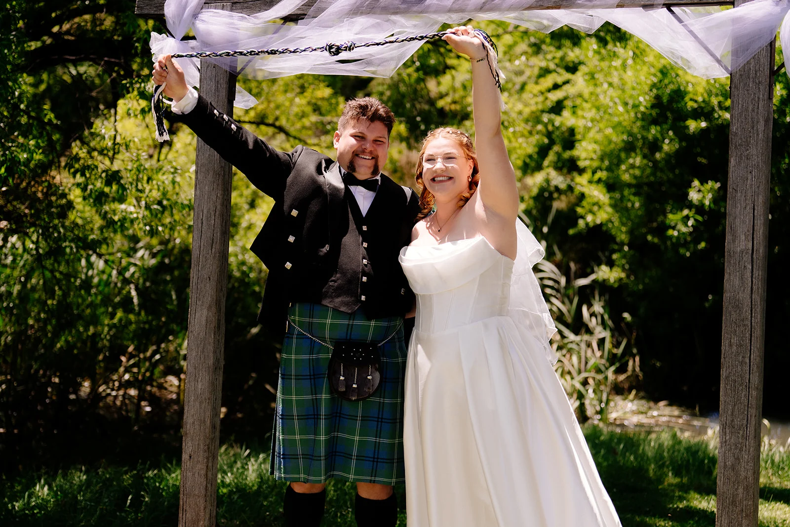 A newly married couple laughing together under a floral arbor, the groom in a tartan kilt