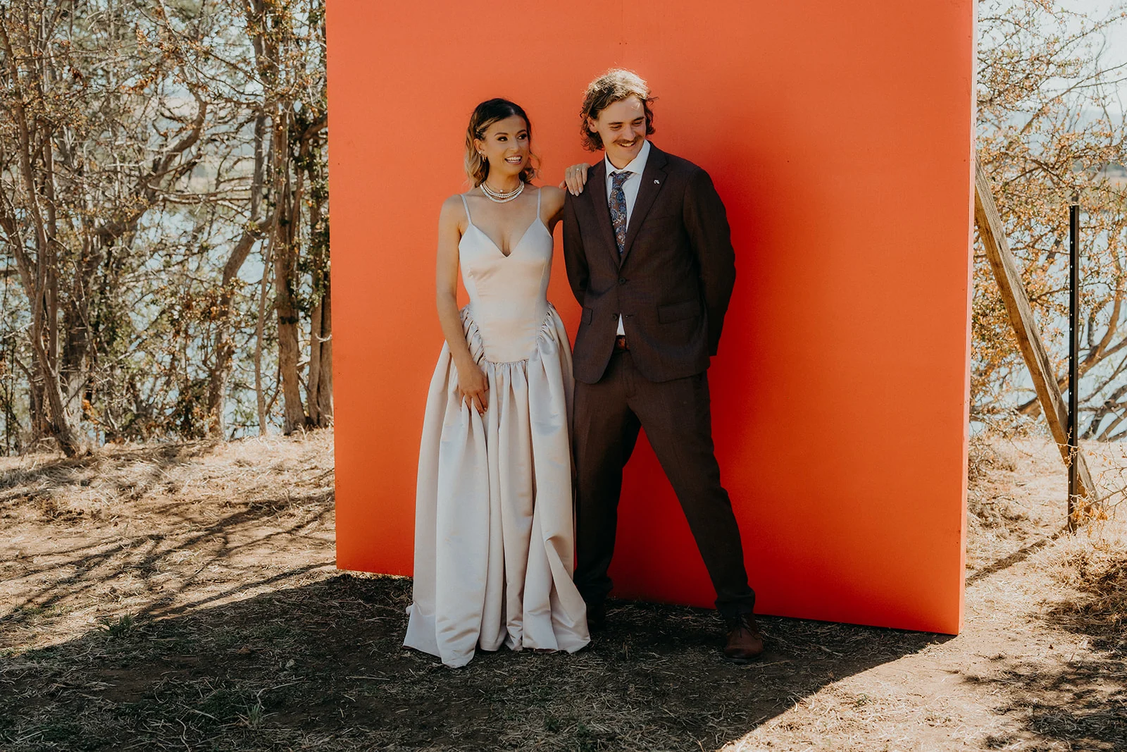 A bride and groom smiling together against a bold orange backdrop