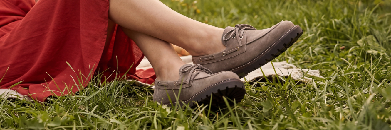 Nahaufnahme der Beine einer Person in einem roten Kleid, die auf einer Decke im Gras sitzt und graue Schnür-Halbschuhe mit robuster Profilsohle trägt.