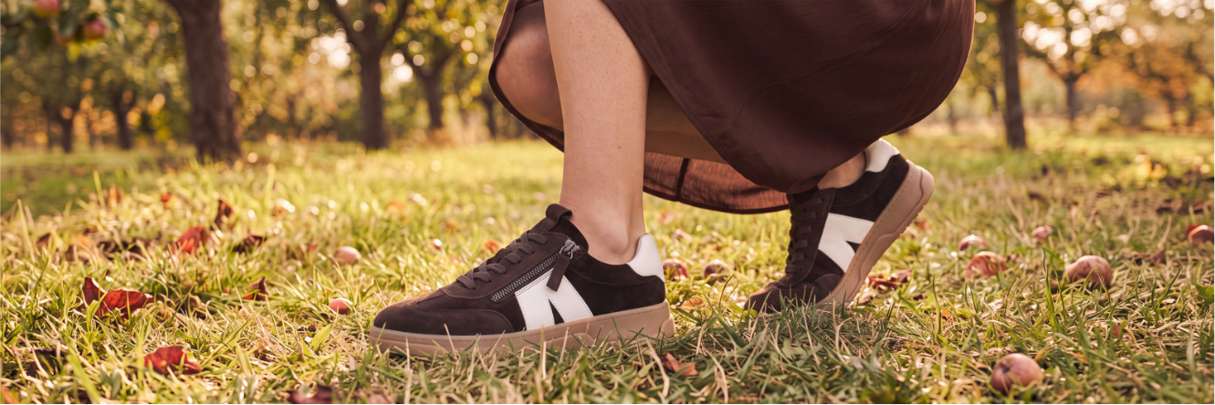 Primo piano delle gambe di una persona con un abito marrone, inginocchiata in un prato di mele e che indossa sneaker marroni con dettaglio bianco e suola chiara, tra l’erba e le mele cadute.