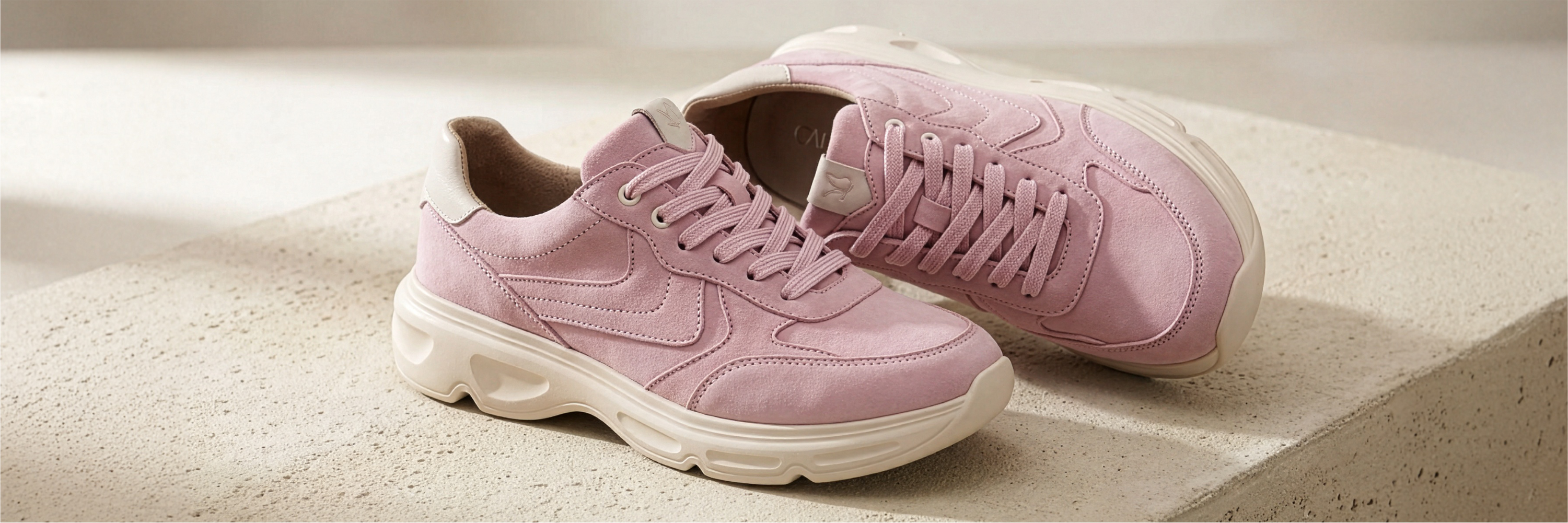 Pink suede sneakers with a white chunky sole on a beige background, one shoe upright, one lying down