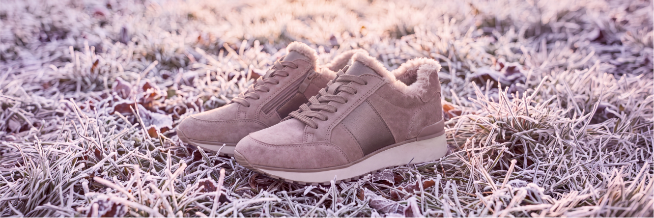 Nahaufnahme eines Paar beigefarbener, gefütterter Winterschuhe im Sneakerstil mit Fellrand, die auf frostbedecktem Gras stehen.