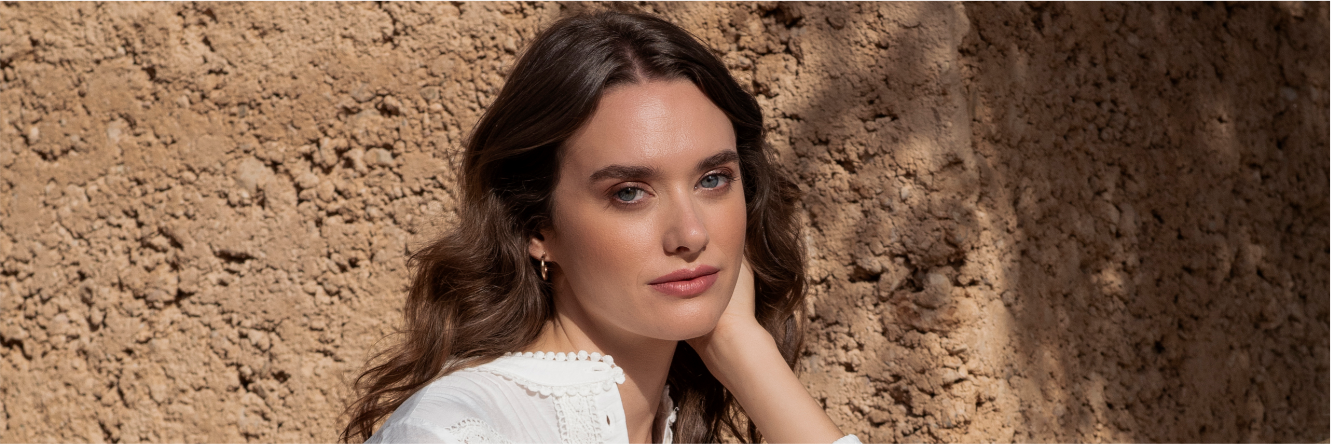 Donna con capelli castani mossi in camicetta bianca davanti a un muro di argilla beige, mano sul viso, trucco naturale