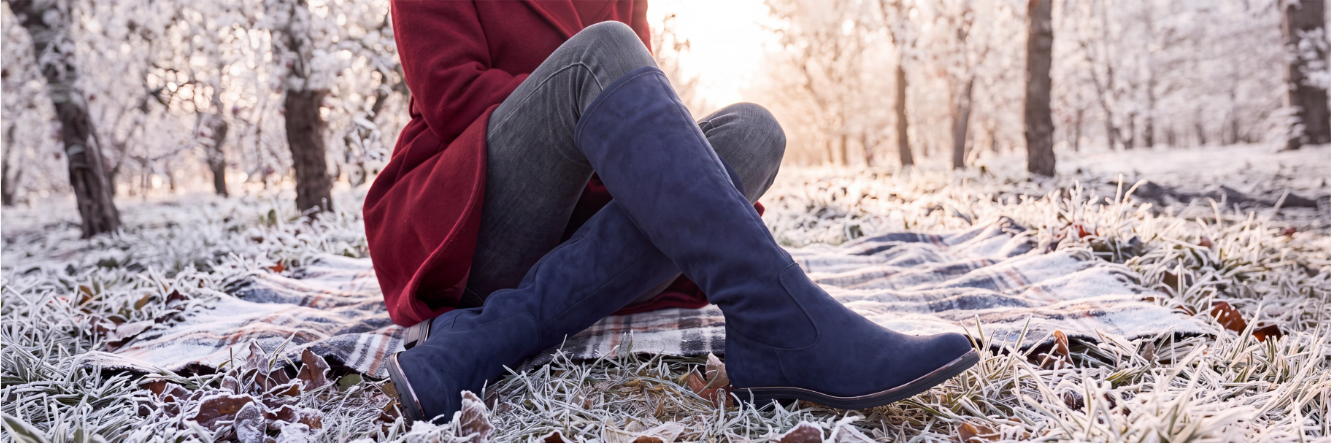 Nahaufnahme einer sitzenden Person auf einer Decke in einer frostigen Winterlandschaft, die blaue kniehohe Stiefel und graue Jeans unter einem roten Mantel trägt.