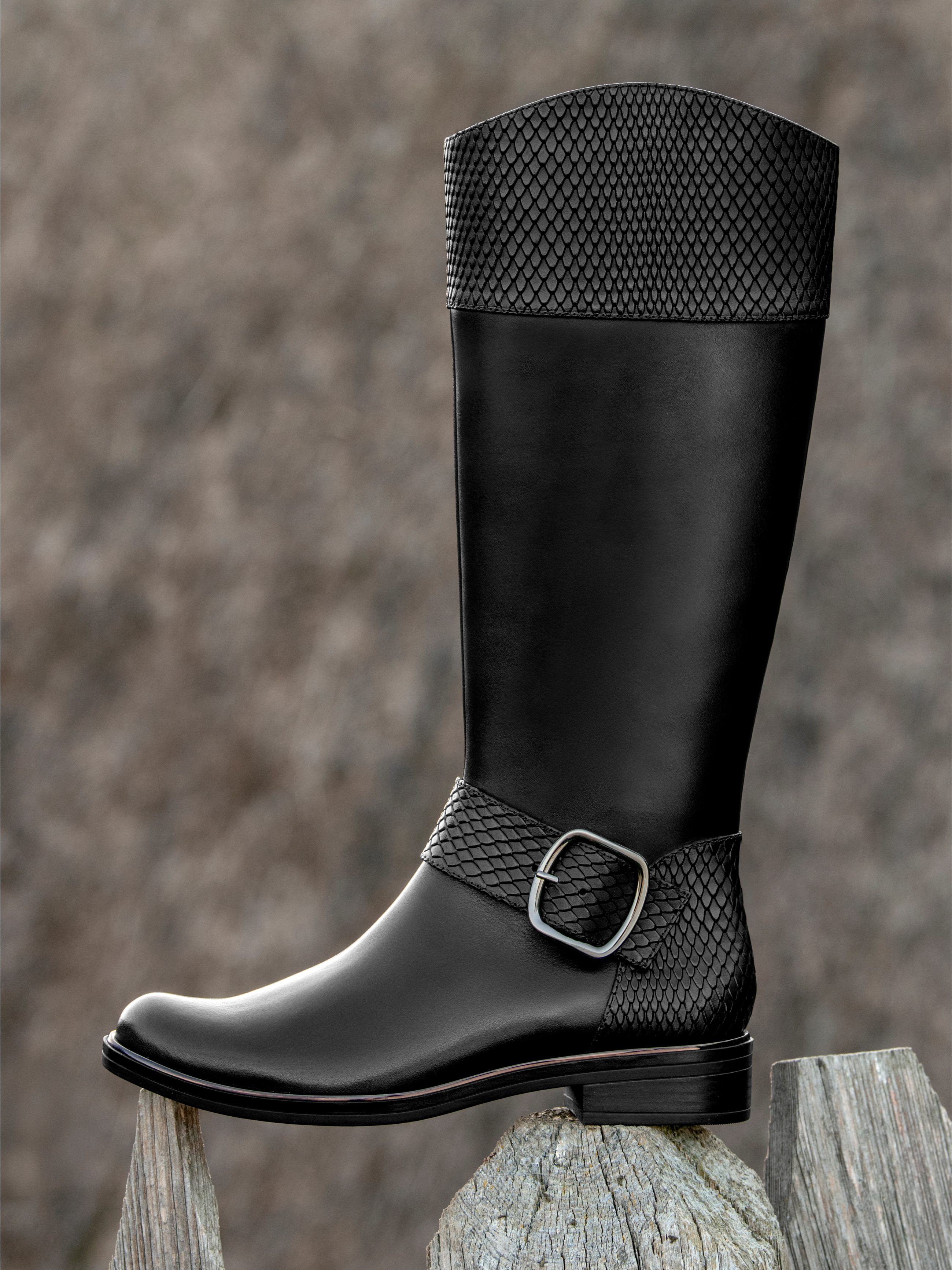 Schwarzer Damenstiefel aus Leder mit Schlangenprägung und Schnalle, auf Holzpfosten vor grauem Hintergrund präsentiert