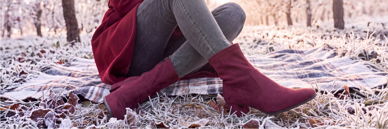 Nahaufnahme einer sitzenden Person auf einer Decke in einer frostigen Winterlandschaft, die bordeauxrote Wildleder-Stiefeletten mit kleinem Absatz und graue Jeans trägt.