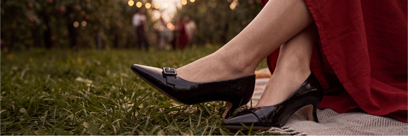 Close-up of a person’s legs in a red dress, sitting on a blanket in the grass and wearing black pointed-toe pumps with a low heel and buckle; in the blurred background, people and string lights in an orchard.