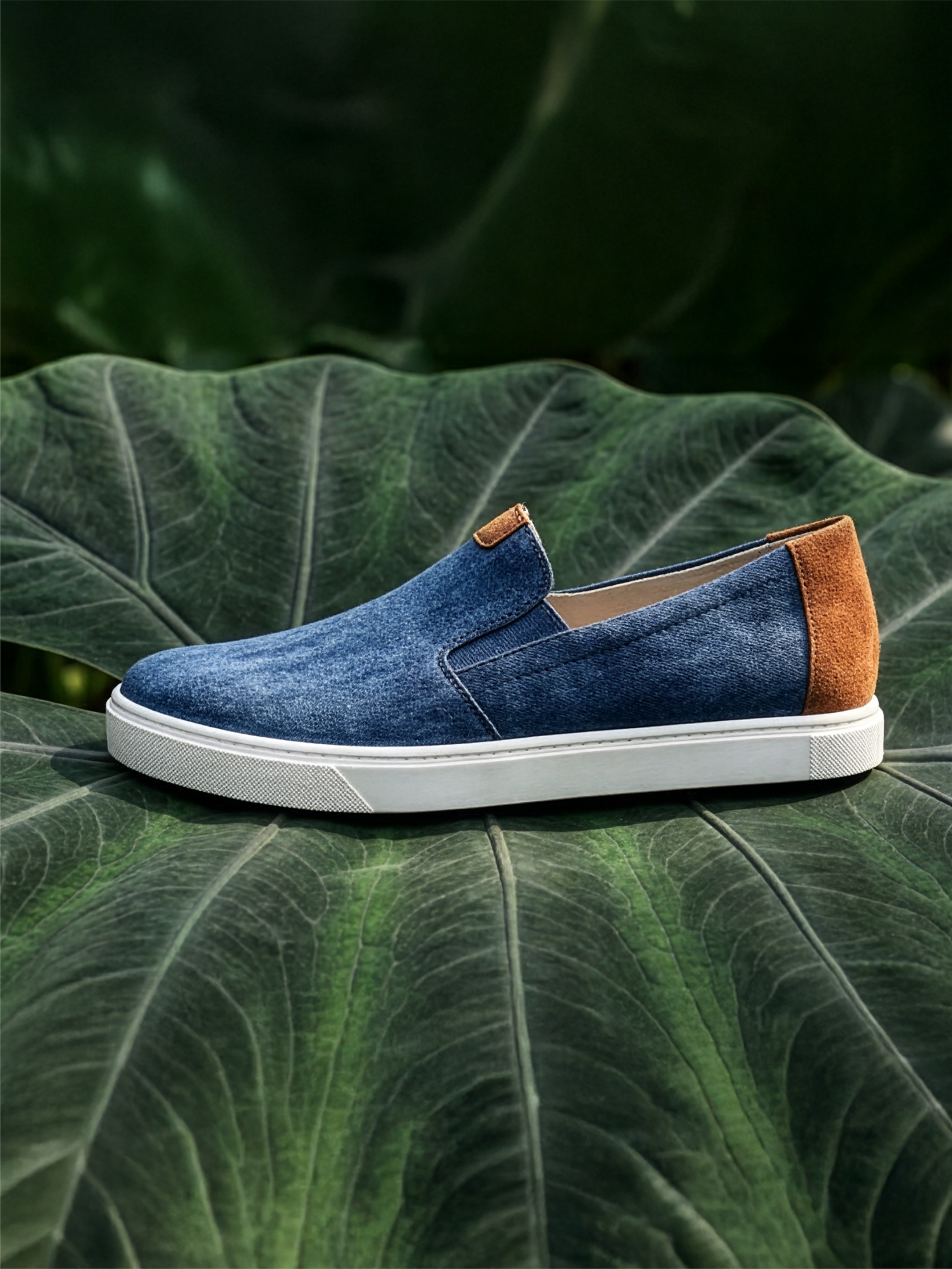 Blue slip-on shoe with brown accents on a white sole, presented on a large green leaf