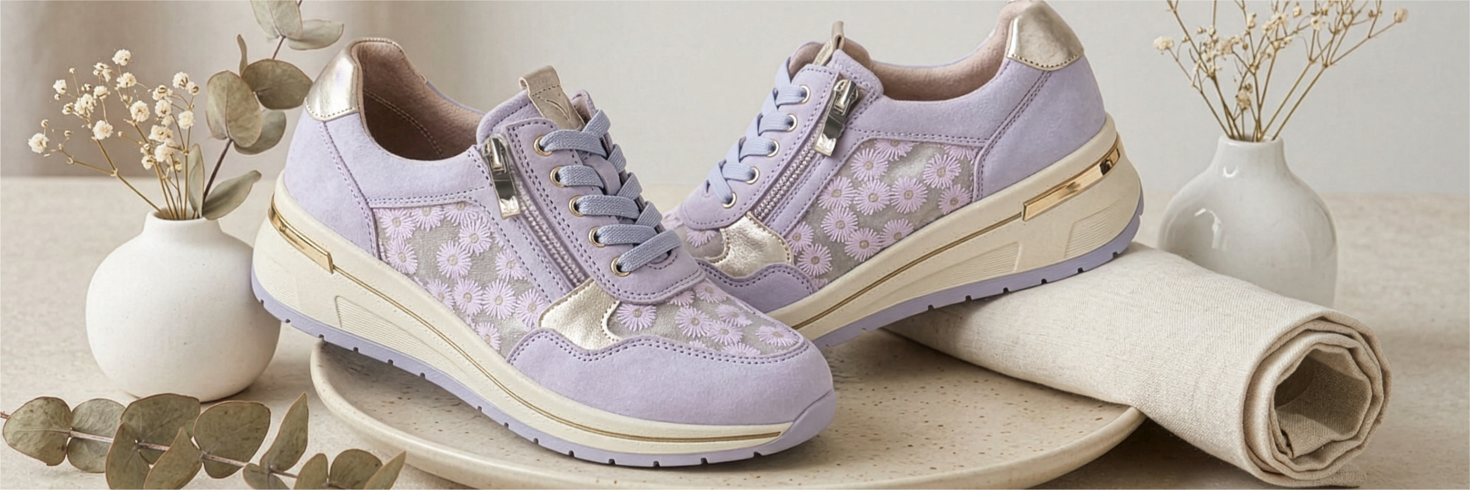 Purple sneakers with floral embroidery and beige soles, decoratively arranged with white vases and dried flowers