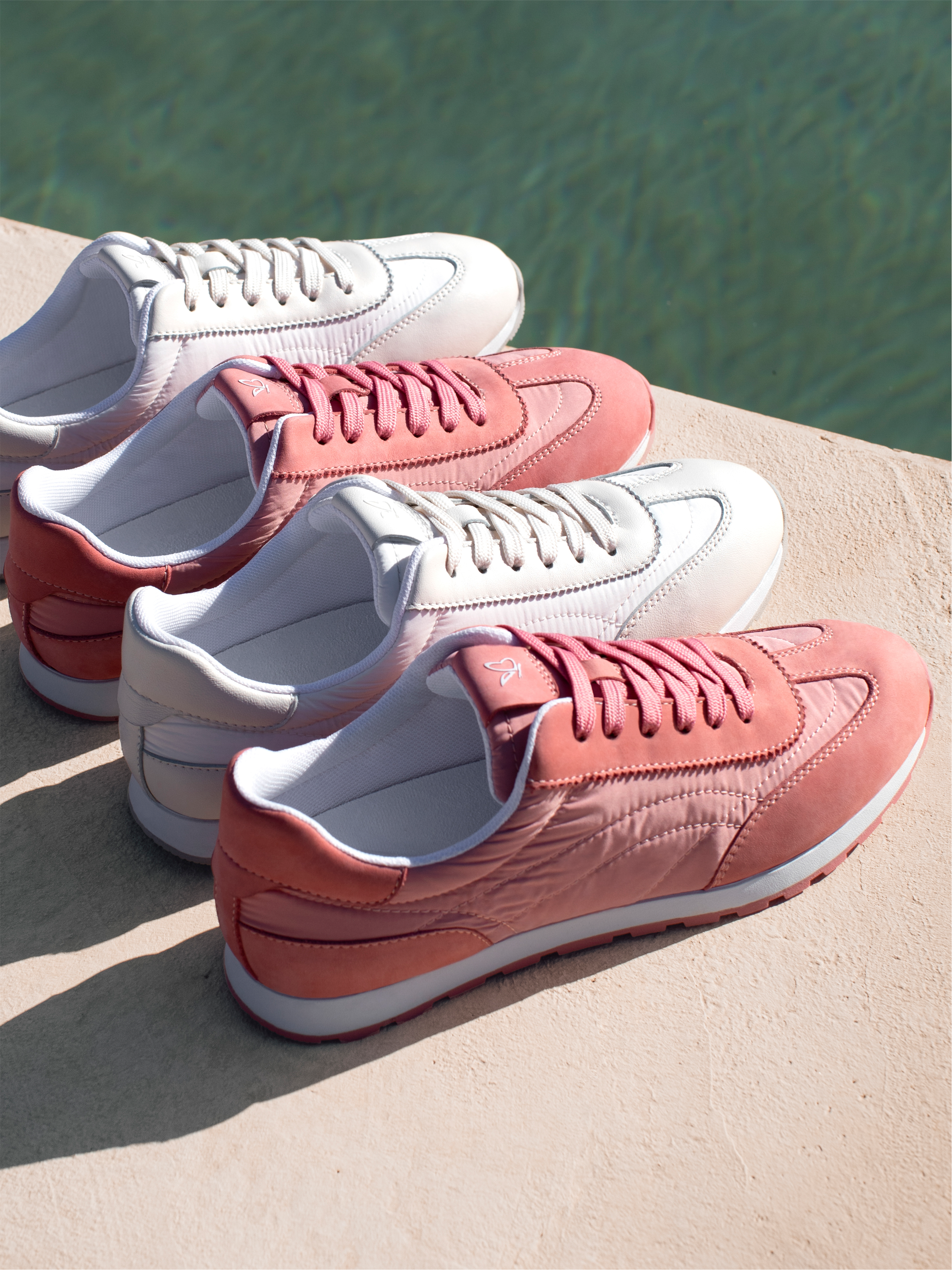 Quattro scarpe da ginnastica in rosa e bianco si trovano una accanto all'altra su un muro di cemento di fronte a un'acqua verde alla luce del sole.