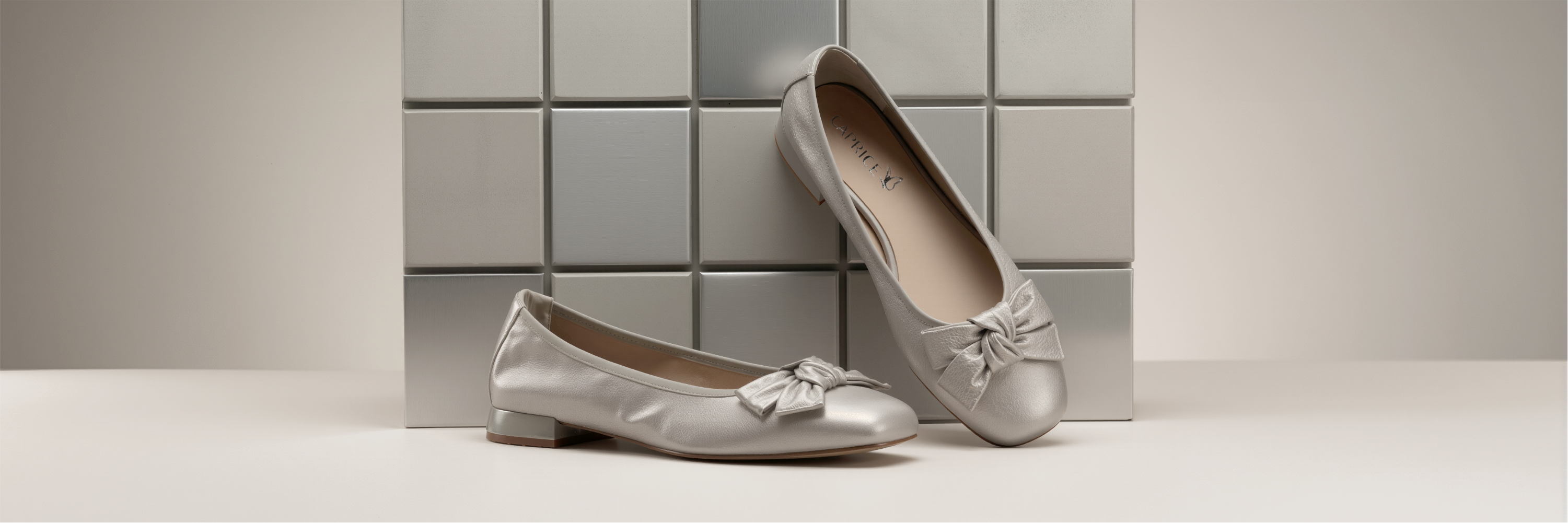 Ballerines argentées pour femmes avec détail de nœud devant un mur de carrelage gris sur fond beige
