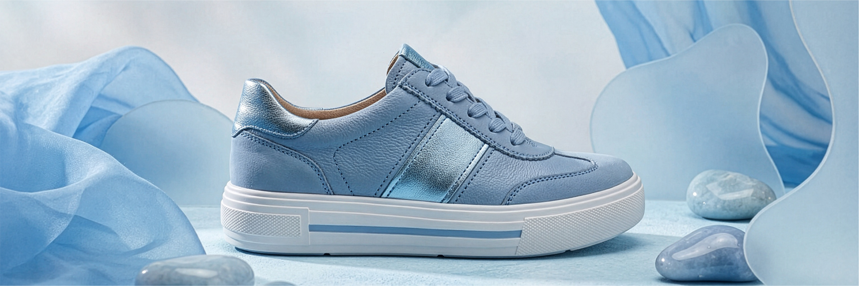 Sneaker in pelle blu con suola bianca, circondata da pannelli in tessuto blu e pietre decorative su sfondo blu.