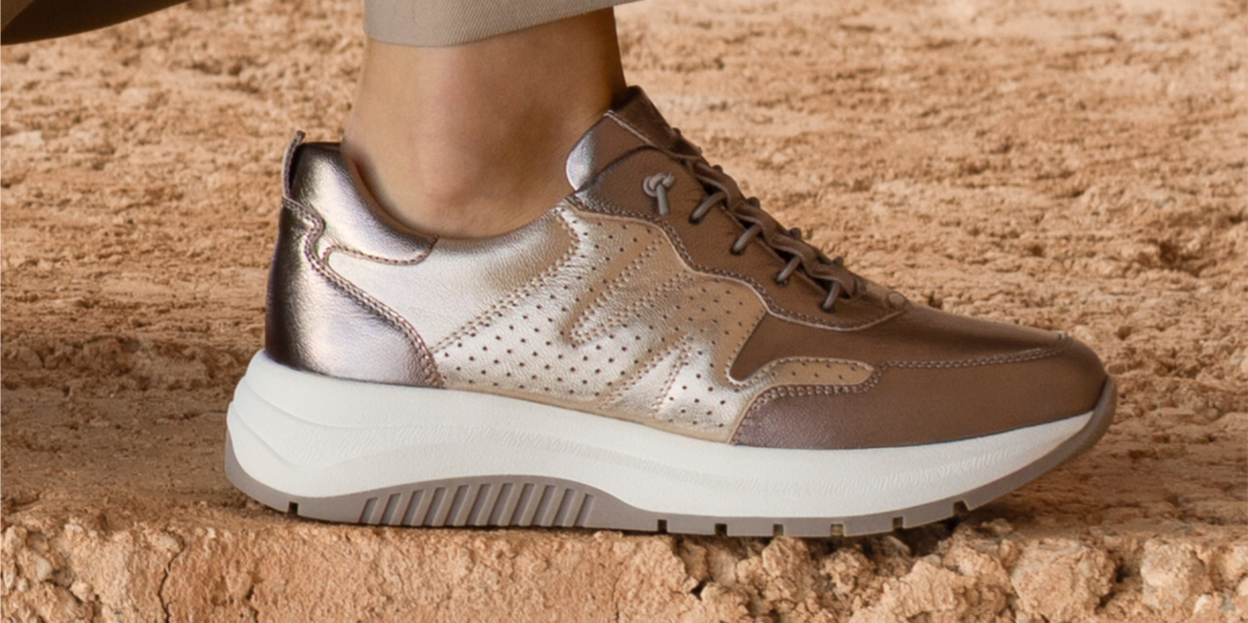 Sneaker in pelle moderne marroni con dettagli metallici dorati appoggiate su un muro di pietra rossa.
