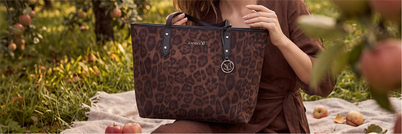 Close-up of a woman sitting on a blanket in an orchard, holding a brown CAPRICE handbag with a leopard print and round charm, surrounded by apples in the grass.