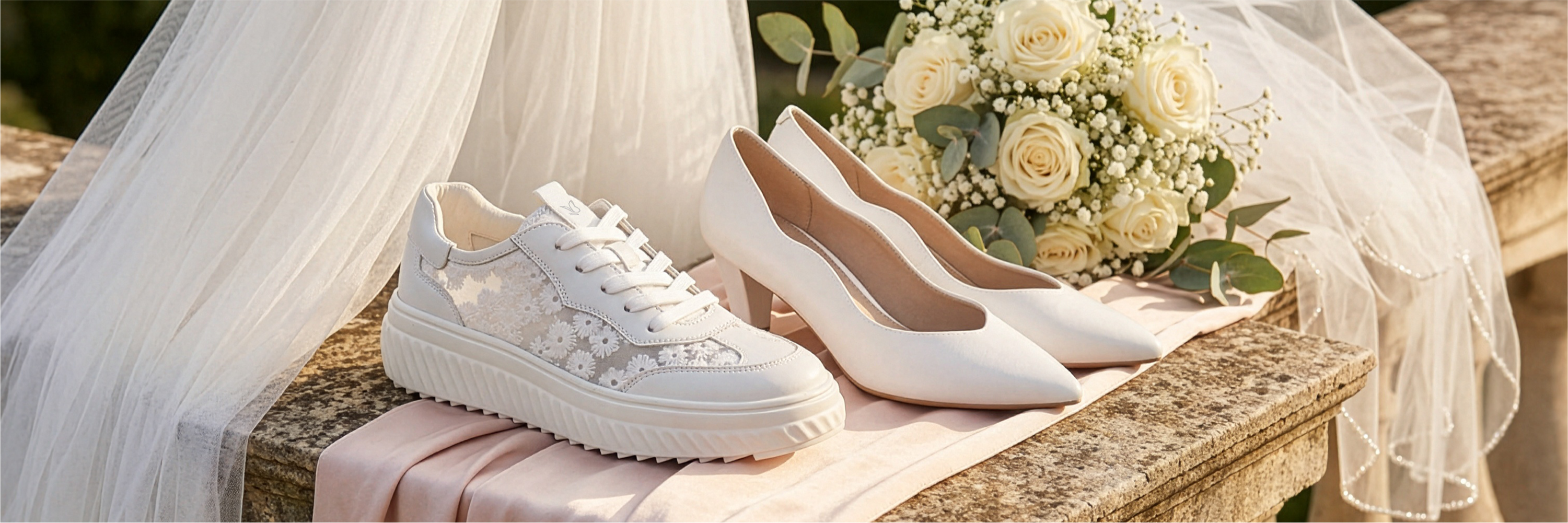 Scarpe da sposa e scarpe da ginnastica bianche su un muro di pietra con un bouquet di rose color crema e velo da sposa sullo sfondo