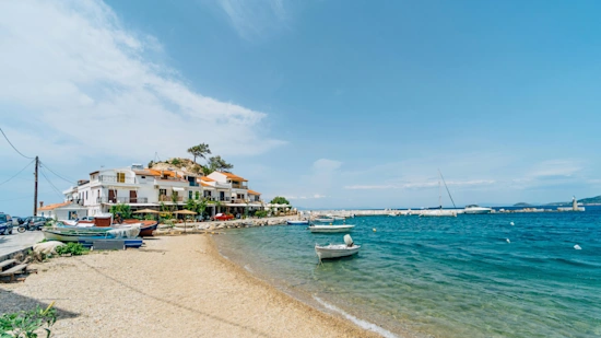 A picture of a coast/ beach in Samos, which a white building in the back ground and boats sailing along the sea.