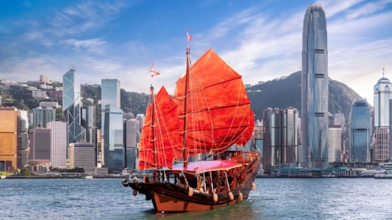 A picture of the Hong Kong harbour, with a red sailboat crossing the Victoria Harbour between Hong Kong Island and Kowloon