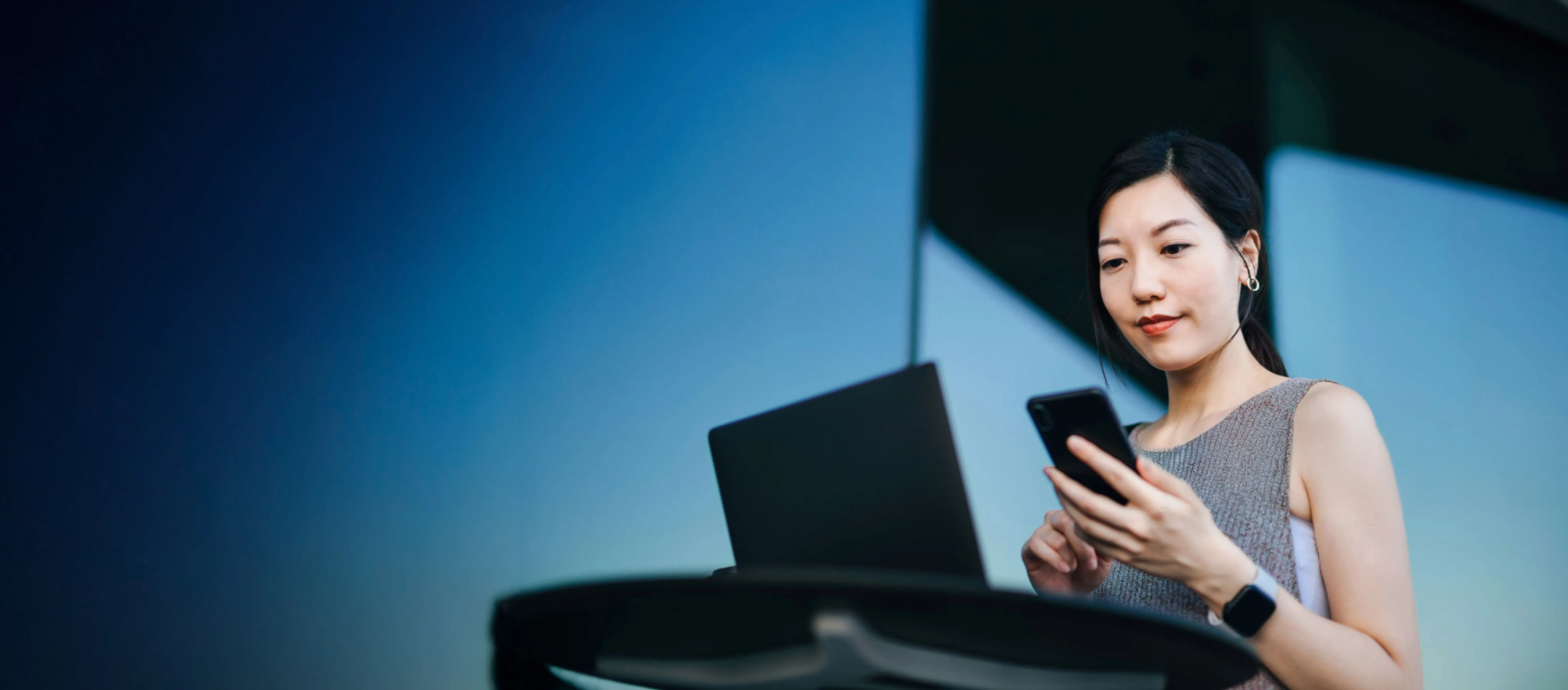 Woman holding a smartphone while standing at a desk with an open laptop, wearing a smartwatch, against a blue background.