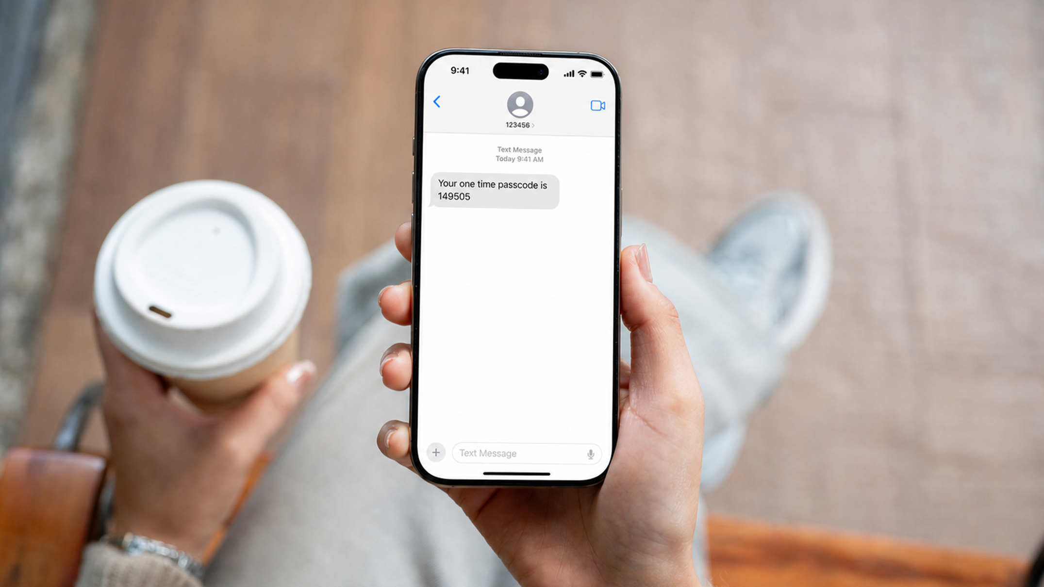 Person holding a smartphone displaying an SMS text message reading "Your one time passcode is 149505," with a coffee cup in the other hand.
