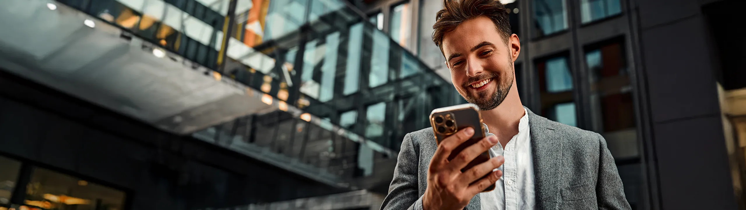 Smiling businessman in a gray blazer looking at his smartphone outside a modern glass office building.