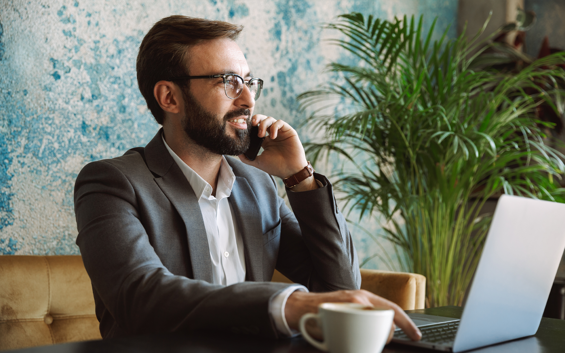 Smiling bearded businessman in glasses talking on phone at a cafe table with a laptop and coffee cup nearby.