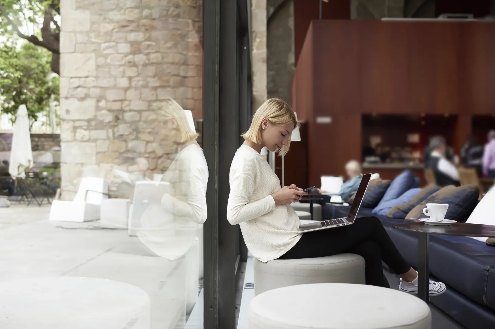 Blonde woman sitting by a cafe window, using a smartphone with a laptop on her lap, reflected in the glass.